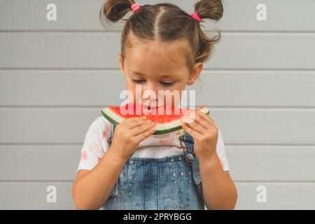 Konzentriertes kleines Mädchen 3-4 Jahre alt mit zwei Pferdeschwänzen isst Wassermelone. Kind im Freien vor dem Hintergrund des Gebäudes hält süße gesunde Früchte in Stockfoto