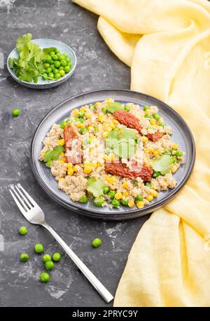 Quinoa-Haferbrei mit grüner Erbse, Mais und getrockneten Tomaten auf Keramikplatte auf grauem Betongrund und gelbem Textil. Seitenansicht, Nahaufnahme. Stockfoto