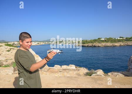 Nahaufnahme, glücklicher Teenager, der sich auf den Drohnenflug am sonnigen Tag vorbereitet, mit dem Meer im Hintergrund Spanien, Balearen, Stockfoto