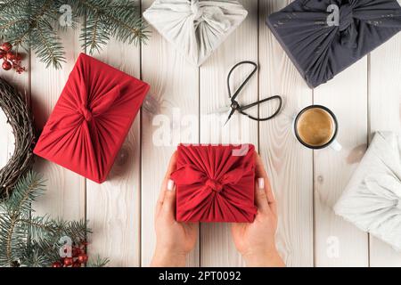 Weibliche Hände halten weihnachtsgeschenk in furoshiki-Paket, Draufsicht. Fichtenzweige, Scheren, Tasse Kaffee und Geschenkschachteln auf Beplankung. Weihnachten oder Stockfoto