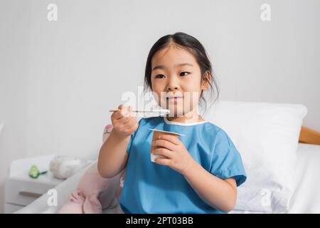 Positives asiatisches Kind im Krankenhauskleid essen köstlichen Joghurt in der Klinik, Stock Image Stockfoto