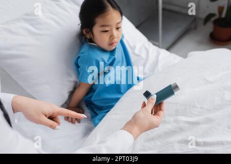 Arzt zeigt auf Inhalator nahe entmutigt asiatisches Mädchen auf Krankenhausbett sitzen, Stock Image Stockfoto