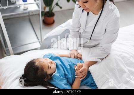 asiatischer Arzt in weißem Kittel, der Hände eines kleinen Patienten hält, der in der Kinderklinik auf dem Bett liegt, Stockbild Stockfoto