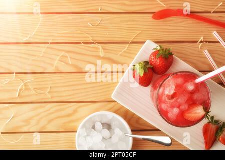 Ein Glas frisch gepflückten, reifen Erdbeersaft mit Eis auf einem Holztisch und einem Behälter mit zerstoßenem Eis. Draufsicht. Horizontale Zusammensetzung. Stockfoto