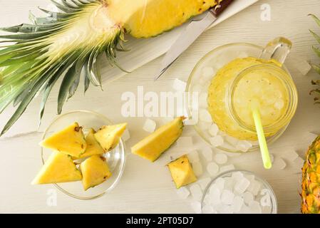 Frisch zubereitetes Ananas-Erfrischungsgetränk mit viel Eis auf einer hölzernen Küchenbank mit Obst und einer Schüssel mit Eis drum herum. Draufsicht. Horizontale Kombi Stockfoto