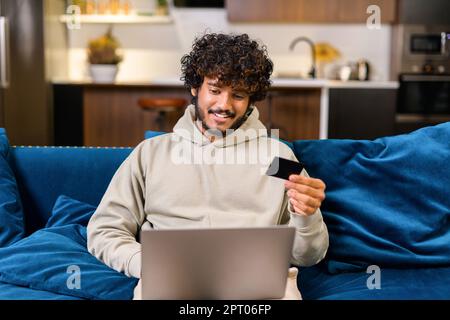 Glücklicher junger, bärtiger Indianer 20s, der zu Hause auf dem Sofa saß, einen Laptop-pc benutzte und eine Kreditkarte hielt, online einkaufte, Essen bestellte Stockfoto