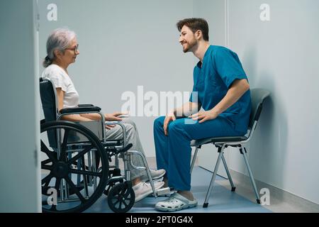 Der Mann in der medizinischen Uniform kommuniziert mit dem älteren Patienten im Rollstuhl Stockfoto