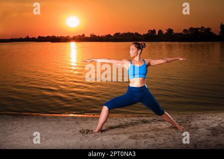 Yoga bei Sonnenuntergang am Strand. Frau, die Yoga macht, Asanas vorführt und das Leben auf dem Fluss genießt Stockfoto