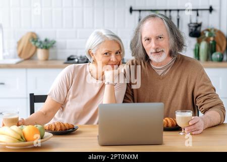 Glückliches, weißes Paar mittleren Alters, Mann und Frau, sitzen zu Hause in der Küche, trinken Croissants und Kaffee zum Frühstück, benutzen Sie einen Laptop zum Frühstück, schauen Sie in die Kamera, lächeln Stockfoto