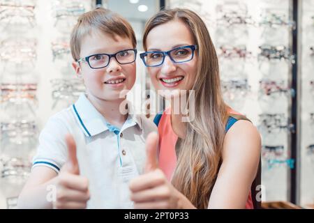 Mutter und Sohn empfehlen Brillen im Optikerladen und Daumen nach oben geben Stockfoto