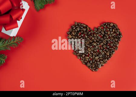 Braune Kaffeebohnen in Herzform mit Christbaumzweigen und Geschenkkarton auf rotem Hintergrund. Flach liegend Stockfoto