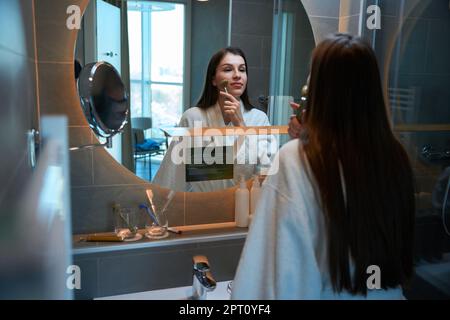 Die langhaarige junge Brünette steht vor dem Spiegel im Badezimmer und gibt sich selbst eine Gesichtsmassage mit einem Jaderoller Stockfoto