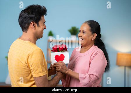 Happy Indian son Wishing Mother day by give greeting Card with flowers to older Mother at Home - Konzept von Beziehung, Bindung und Genuss Stockfoto