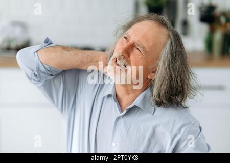 Nahaufnahme Foto eines ungesunden weißen älteren grauhaarigen Mannes, sitzt in der Küche am Tisch, fühlt sich überarbeitet, müde, hat Schmerzen im Nacken, massiert seinen Hals mit seinen Händen, Augen geschlossen, grimmig Stockfoto