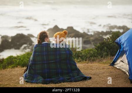 Es wird gemütlich. Rückansicht eines anhänglichen jungen Paares, das in eine Decke gehüllt wurde, auf ihrem Campingplatz Stockfoto