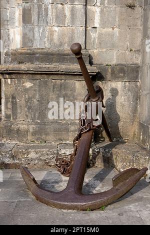 Großer antiker Schiffsanker, der sich an die Wand lehnt. Steinmauer, Mauerwerk, Textur, Bronzefarbe, Schatten, Usw. Stockfoto