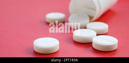 Mehrere weiße Tabletten fallen auf der roten Oberfläche aus dem Kunststoffbehälter. Hintergrundbild zu medizinischen und pharmazeutischen Themen Stockfoto