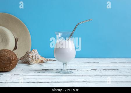 Ein Glas Kokosmilch-Cocktail mit wiederverwendbarem Metallcocktail, eine Kokosnuss, einen Sommerhut und eine exotische Muschel auf einem weißen Holztisch mit Kopie s Stockfoto