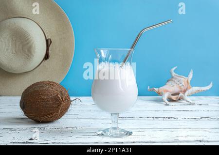 Ein Glas Kokosmilch-Cocktail mit wiederverwendbarer Metallsonde, eine Kokosnuss, einen Sommerhut und eine exotische Muschel auf einem weißen Holztisch, Sommerorganisation Stockfoto