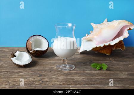 Ein Glas Kokosmilch-Cocktail, eine Kokosnuss und eine exotische Muschel auf einem Holztisch, Sommergetränk aus biologischem Anbau Stockfoto