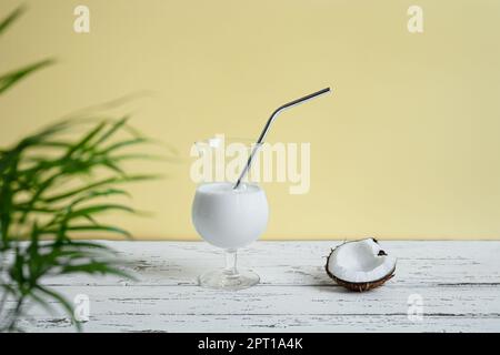 Ein Glas Kokosnuss-Milchshake mit wiederverwendbarer Cocktailröhre aus Metall und ein Stück Kokosnuss auf einem weißen Holztisch Stockfoto