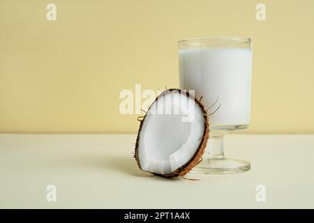 Ein Stück Kokosnuss neben einem Glas vegane Kokosmilch auf hellgelbem Hintergrund Nahaufnahme mit Kopierbereich, alternatives Milchkonzept Stockfoto