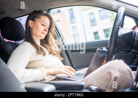 Junge Geschäftsfrau, die an einem Laptop arbeitet, sitzt in einem Auto hinter dem Steuer, Arbeitsplatz im Auto Stockfoto