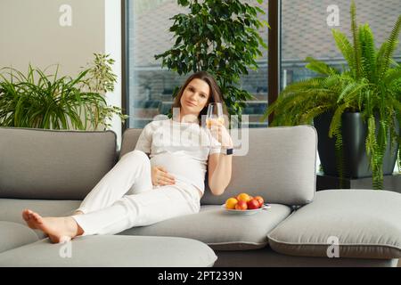 Die süße Schwangere sitzt mit einem Glas frischen Orangensaft in der Hand auf dem Sofa Stockfoto