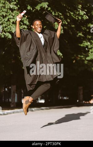 Glücklicher afroamerikanischer Absolvent von der Universität, der mit Glück in schwarzem Mantel und Hut hüpft. Foto eines Studenten an einem sonnigen Tag. Höhere Bildung Stockfoto