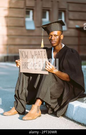 Ein verärgerter Schwarzer sitzt mit einem Plakat auf der Straße und sucht einen Job. Universitäts- oder Universitätsstudenten mit Graduiertenkleid und -Mütze. Bildung Stockfoto