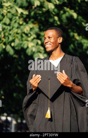 Glücklicher afroamerikanischer Student im Kittel steht an sonnigen Tagen draußen und hält den Abschlussmütze in den Händen. Stellensuche, Hochschulabschluss Stockfoto