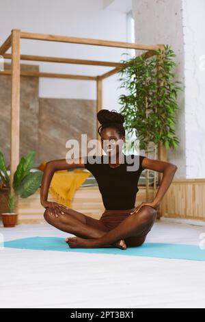 Wunderschöne und sportliche afro-amerikanische Frau, die in Schmetterlingsposition sitzt und Yoga auf einer Turnmatte in einem dekorierten Studio macht. Muskeldehnung, Gymnastik Stockfoto