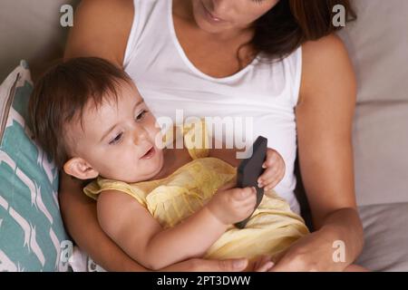 Sie ist so neugierig. Ein kleines Mädchen, das auf dem Schoß seiner Mutter liegt und mit dem Handy spielt Stockfoto