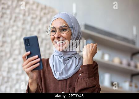 Eine angenehm überraschte junge muslimische Frau in einem Hidschab zu Hause sieht mit Freude am Telefon. Geld empfangen, gute Nachrichten lesen, Nachrichten. Zeigt eine Siegesgeste mit seiner Hand. Stockfoto