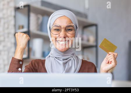 Nahaufnahme. Muslimische junge Frau im Hidschab, die zu Hause sitzt, mit Laptop und Kreditkarte. Glücklich vor der Kamera. Haben einen Traum wahr werden lassen, haben online eingekauft. Stockfoto