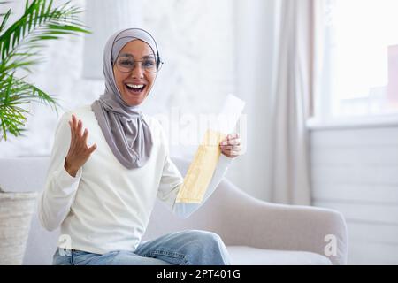 Junge muslimische Studentin in Hijab sitzt zu Hause auf dem Sofa und liest den Brief der Universität mit den Ergebnissen der Prüfung, des Interviews, der Jobannahme. Ich freue mich, in die Kamera zu sehen. Stockfoto