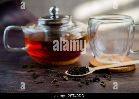 Ein Holzlöffel mit Tee liegt auf einem dunklen Holzhintergrund, ein Becher und eine Teekannen sind von hinten zu sehen, ein Foto in dunklen Farben. Hochwertige Fotos Stockfoto