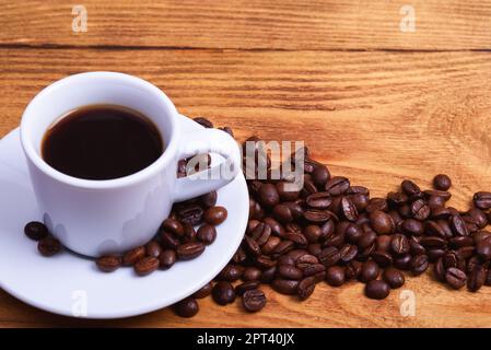 Frisch gemachter Kaffee in einer weißen Tasse auf einer weißen Untertasse, umgeben von verstreuten Kaffeebohnen auf einem Holztisch. Frisch gerösteter Kaffee. Platz für einen Inscri Stockfoto