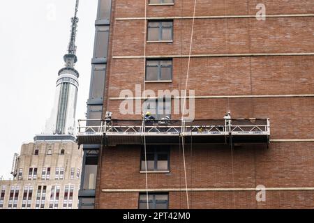 Die Arbeiter führen auf einer abgehängten Plattform Ziegelsteine mit dem Empire State Building im Hintergrund durch, 2023, New York City, USA Stockfoto