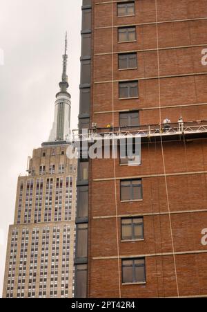 Die Arbeiter führen auf einer abgehängten Plattform Ziegelsteine mit dem Empire State Building im Hintergrund durch, 2023, New York City, USA Stockfoto