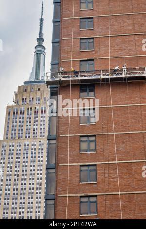 Die Arbeiter führen auf einer abgehängten Plattform Ziegelsteine mit dem Empire State Building im Hintergrund durch, 2023, New York City, USA Stockfoto