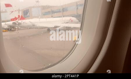 Blick auf den Flughafen durch das Flugzeugfenster, nasses Bullauge, verschwommenes Bild des Flugzeugs, das Konzept des Fliegens bei Regenwetter. Stockfoto