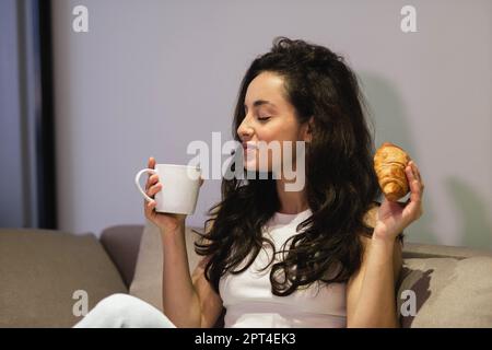 Nahaufnahme einer hübschen weißen jungen Frau mit umstrittenem Gesicht und geschlossenen Augen, die drinnen sitzt und Kaffee mit Croissant zum Frühstück genießt. Junge Frau Stockfoto