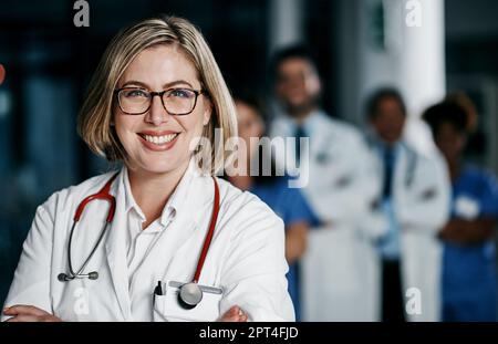 Lernen Sie das Team kennen, das den Unterschied macht. Porträt einer selbstbewussten jungen Ärztin, die im Krankenhaus mit ihren Kollegen im Hintergrund arbeitet Stockfoto