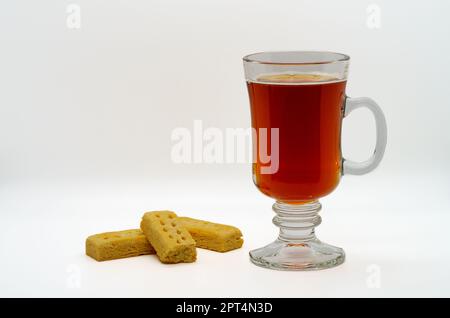 Heißer Tee, der in einer klaren Tasse serviert wird, begleitet von bereits knabberten Shortbread-Snacks Stockfoto