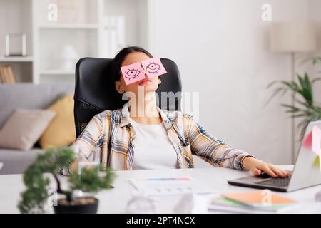 Das Konzept einer Menge Arbeit. Müde indische Studentin, die mit Aufklebern auf den Augen schläft, während sie am Tisch sitzt. Abgelenkte Frau im Heimbüro Stockfoto