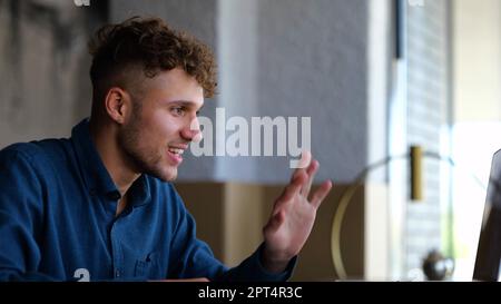 Nahaufnahme eines fröhlichen jungen weißen Mannes, der lächelt und mit der Hand winkt, auf den Laptop-Bildschirm schaut und sich online im Restaurant mit Video chattet. Ein hübscher Kerl Stockfoto