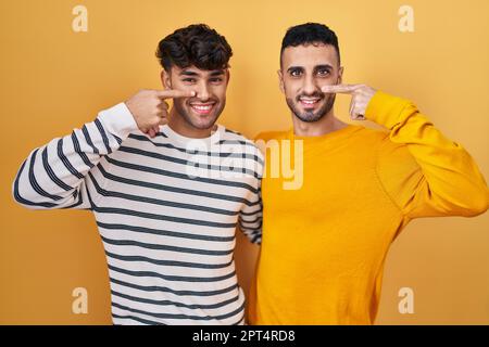 junges lateinamerikanisches schwules Paar, das auf gelbem Hintergrund steht und mit dem Finger an Gesicht und Nase zeigt, fröhlich lächelt. Schönheitskonzept Stockfoto
