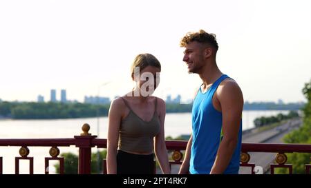 Ein fröhliches, junges Paar von Sportlern, die draußen stehen und miteinander reden und lachen. Porträt von Weißen, die sich nach dem Training auf der Straße unterhalten Stockfoto