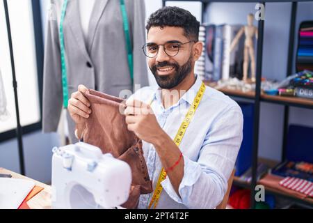 Junger arabischer Mann, Schneider, lächelnd, hält selbstbewusst Jeans in der Schneiderei Stockfoto